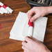 A hand using red self-adhering napkin tape to fold a white napkin.
