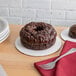 A chocolate cake on a white corrugated cake circle with chocolate chips and drizzle, with a fork on a table.