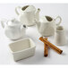 A group of Tuxton eggshell white teapots on a white surface with a bowl of cinnamon sticks.