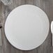 A 10 Strawberry Street white stoneware charger plate on a wood surface with silverware and a glass of water.