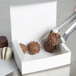 A hand using metal tongs to place a chocolate truffle in a white candy box.