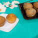 A teal lagoon Creative Converting tissue/poly table cover on a table with a plate of muffins and a plate of cupcakes.