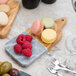 An American Metalcraft olive wood and marble serving board with a plate of macarons on a white surface.