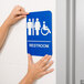 A person holding a blue and white Tablecraft ADA restroom sign with braille and symbols.