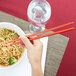 A hand holding red plastic Town chopsticks over a bowl of noodles.