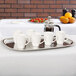 A Vollrath stainless steel oblong serving tray with white coffee cups and a coffee pot on a table.