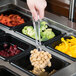 A person using a Cambro clear perforated salad bar spoon to serve food into a container.