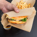 A hand holding a sandwich in a Bagcraft Packaging EcoCraft brown paper cone bag with a burger and fries inside.