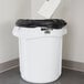 A hand throws a piece of paper into a white Rubbermaid BRUTE trash can.