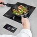 A hand using a Vollrath induction cooker to cook vegetables in a pan on a countertop.