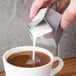 A person pouring milk from an American Metalcraft stainless steel milk carton creamer into a cup of coffee.