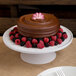 A chocolate cake with berries on top sits on a faux Carrara marble round plate stand on a table in a bakery display.