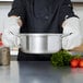 A chef wearing gloves holding a Vollrath stainless steel sauce pot.