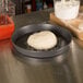 A dough ball in an American Metalcraft hard coat anodized aluminum pizza pan on a counter.