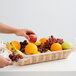 A hand reaching for an apple in a Tablecraft rectangular woven rattan-like basket full of fruit.