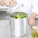 A person using a Vollrath aluminum steamer to cook broccoli with a bowl of peas in it.