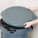 A person's hand reaching for a grey Rubbermaid trash can lid.