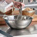A person pouring milk into a Vollrath stainless steel mixing bowl.