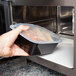 A hand holding a Pactiv Newspring VERSAtainer square black plastic container of food in a microwave.