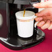 A hand pours brown coffee from a 2.5 Liter Gravity Container into a white cup.