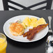 A Tuxton bright white china plate with bacon, eggs, and a biscuit on a table.