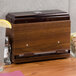A dark walnut woodgrain Vollrath Straw Boss dispenser on a counter with a glass of water next to it.