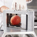 A gloved hand uses a Vollrath scalloped blade assembly to slice a tomato in a machine.