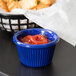 A navy blue fluted melamine ramekin filled with ketchup on a table with a basket of french fries.