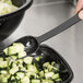 A hand using a black Thunder Group perforated salad bar spoon to scoop cucumbers from a bowl.