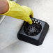 A person in a yellow glove holding a black square plastic floor drain strainer with holes.