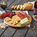 A Tablecraft acacia wood round display board with cheese, grapes, and bread on a table.