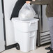 A person putting a plastic bag into a white Rubbermaid wheeled trash can.