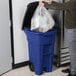 A person putting a plastic bag into a blue Rubbermaid trash can.