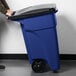 A person pushing a Rubbermaid blue wheeled rectangular trash can with a black lid.
