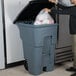 A man putting a plastic bag into a Rubbermaid grey wheeled trash can.