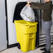 A man putting a plastic bag of trash into a yellow Rubbermaid trash can.