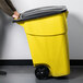 A person pushing a Rubbermaid yellow wheeled rectangular trash can with a black lid.