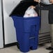 A man putting a plastic bag in a blue Rubbermaid rectangular trash can.