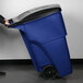 A person pushing a Rubbermaid blue wheeled rectangular trash can with a lid.