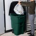 A man putting a plastic bag into a Rubbermaid green wheeled trash can.