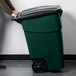 A man pushing a green Rubbermaid wheeled rectangular trash can.