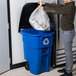 A man putting a plastic bag into a blue Rubbermaid recycling bin.