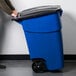 A person pushing a Rubbermaid blue wheeled recycling bin.