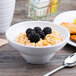 A white Elite Global Solutions melamine bowl filled with oatmeal and blackberries with a spoon on the table.