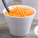 A white melamine crock filled with shredded carrots with a spoon in it.