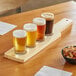 An Acopa wooden flight paddle with four pub tasting glasses of beer on a table with a bowl of mixed nuts.