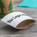 A white customizable cardboard coffee cup sleeve with black text on a table with a white coffee cup.