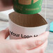 A hand holding a white coffee cup with a green and white customizable sleeve.