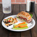A white Elite Global Solutions 3-compartment melamine plate with a sandwich, carrots, and a glass of liquid on a table.