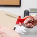 A hand holding 3M Scotch multi-purpose scissors with red and gray handles cutting a piece of paper.